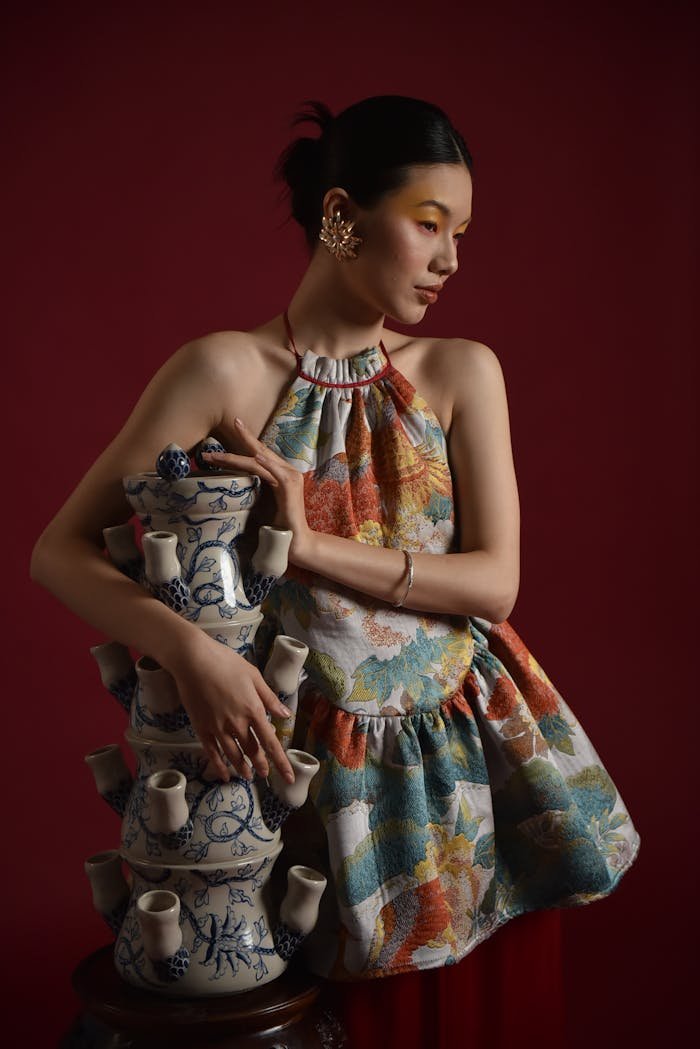 An elegant woman in a floral dress holding an intricate ceramic sculpture, set against a deep red backdrop.